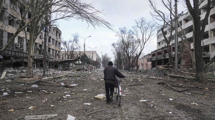 A cyclist walks down a damaged road with destroyed buildings on either side